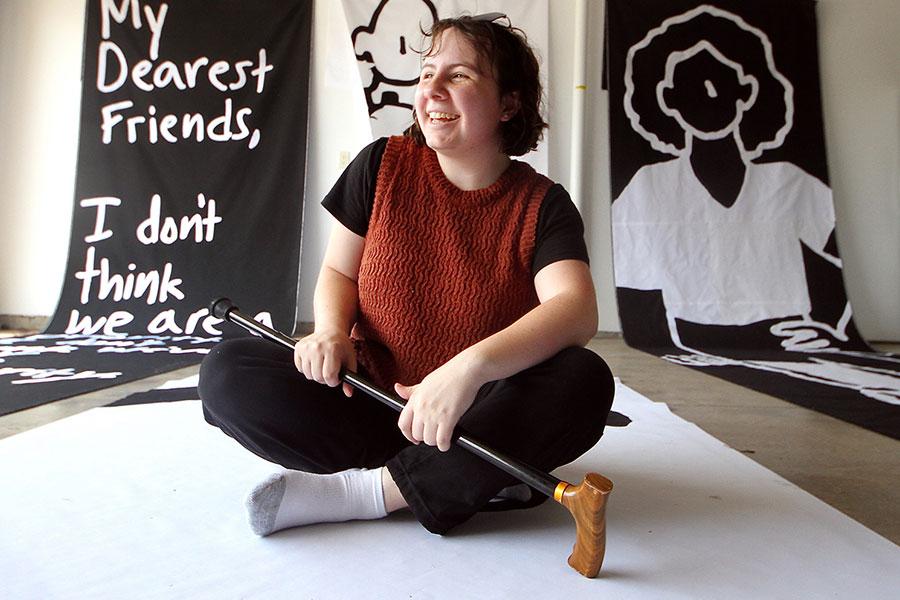 Oaklee Thiele, a petite dark-haired woman, smiles as she sits cross-legged in front of several large, black-and-white banners.