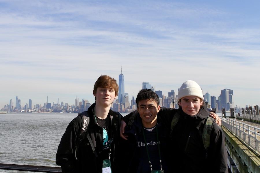 Three male students dressed in warm coats stand by the water, with the New York skyline behind them.