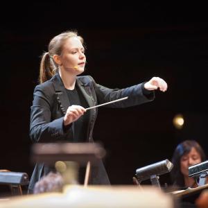 A blonde-haired female conductor leads an orchestra.
