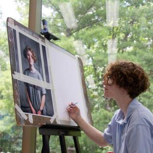 A student in a light blue shirt works on a self-portrait beside a window looking out into the forest.
