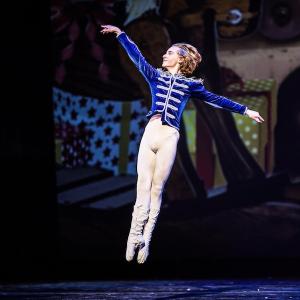 A dancer dressed in a blue jacket and white tights leaps into the air.
