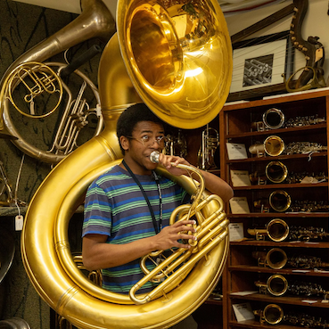 Person playing an early sousaphone with shelves of brass and woodwind instruments behind.