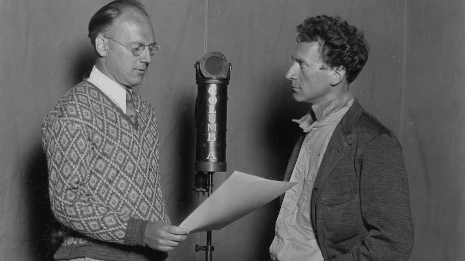 Black and white photo of two men speaking into a vintage "COLUMBIA" microphone.