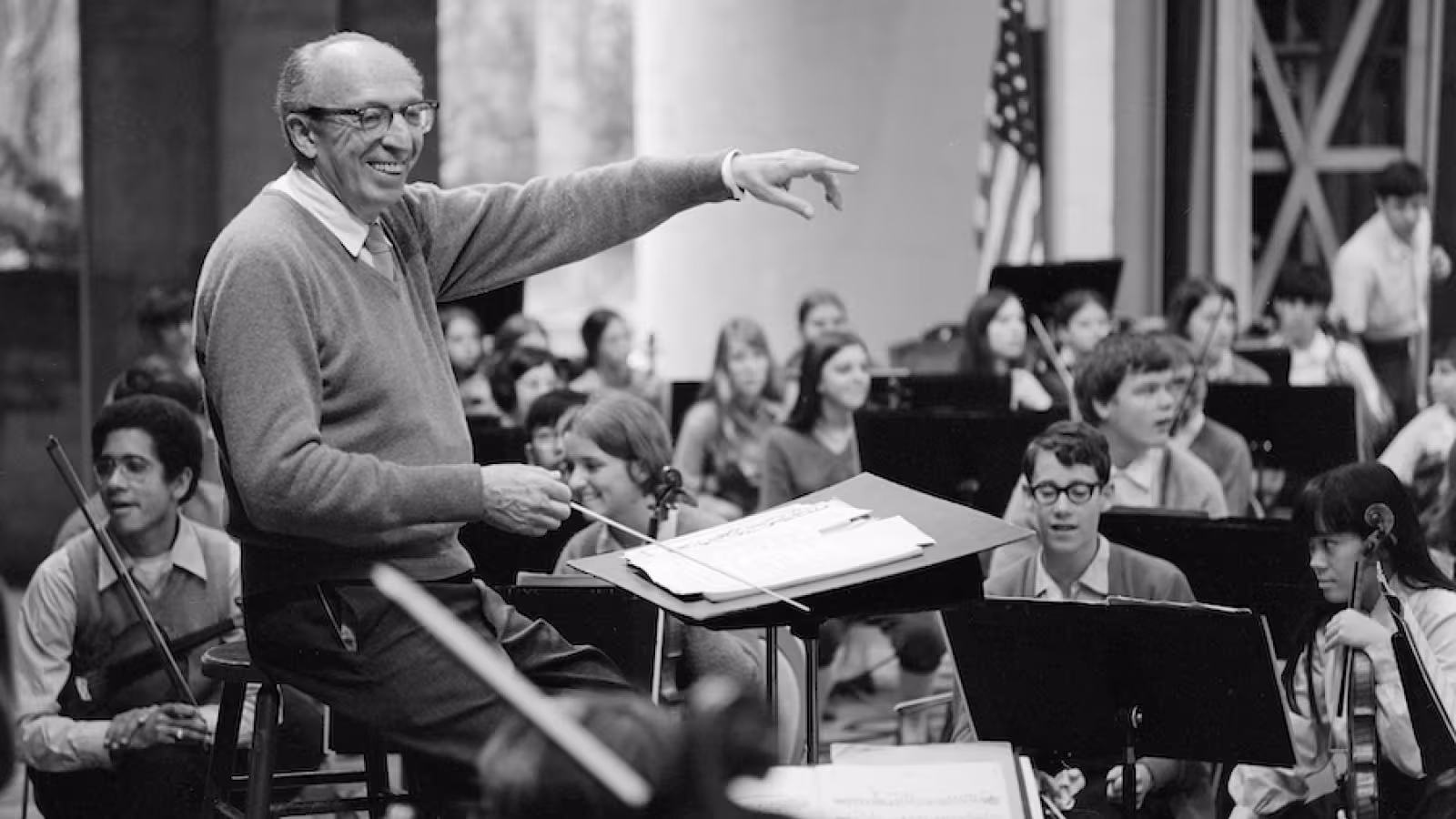 An conductor leads a youth orchestra, with musicians holding string instruments.