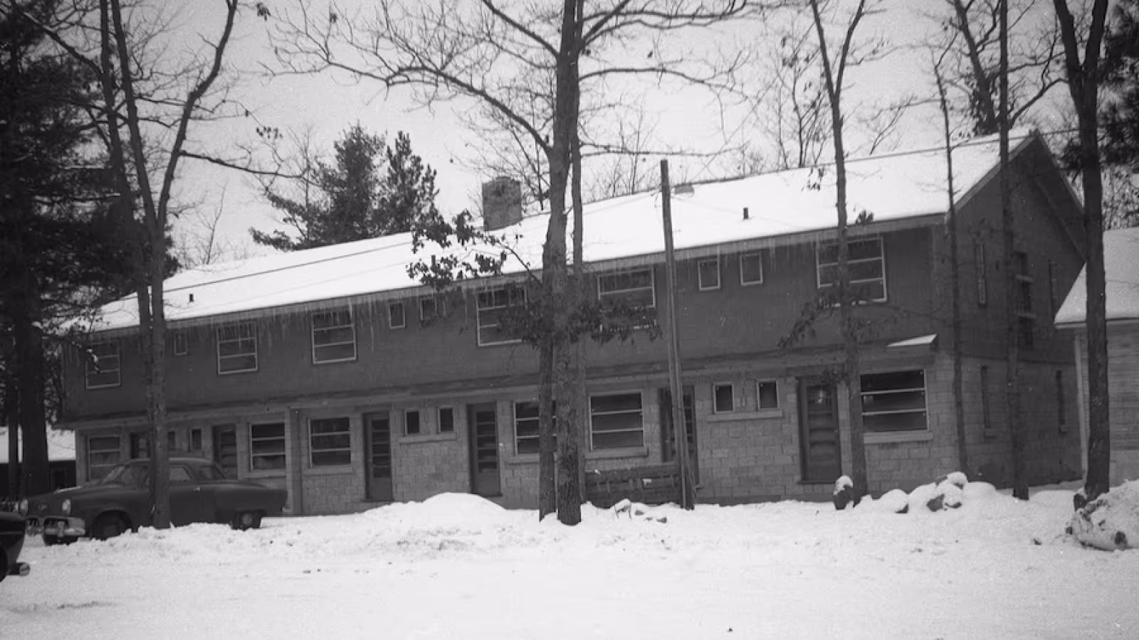 Black and whoite photo of a snow-covered two-story building with icicles on the roof and bare trees around.