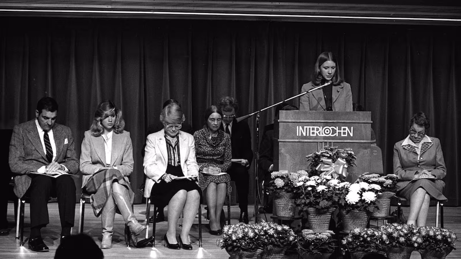 A group of people on stage, with a woman speaking at a podium labeled "INTERLOCHEN," surrounded by seated individuals and floral arrangements.