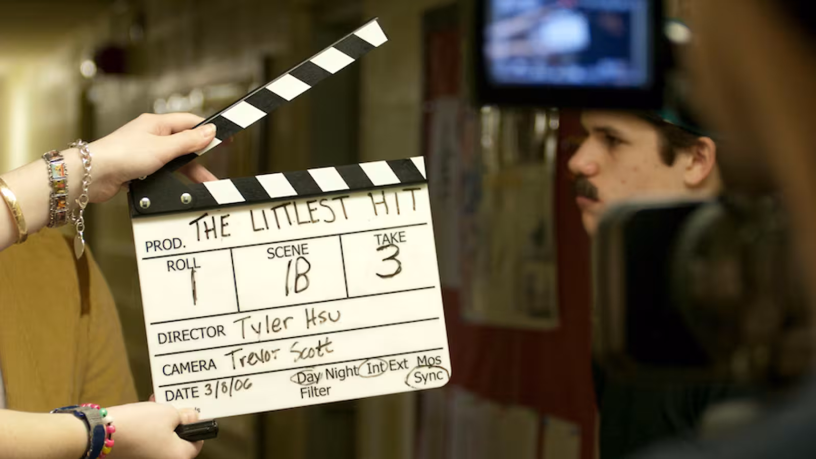 A clapboard in a film scene setup with production details, held in the foreground, while a person is recorded in the background.
