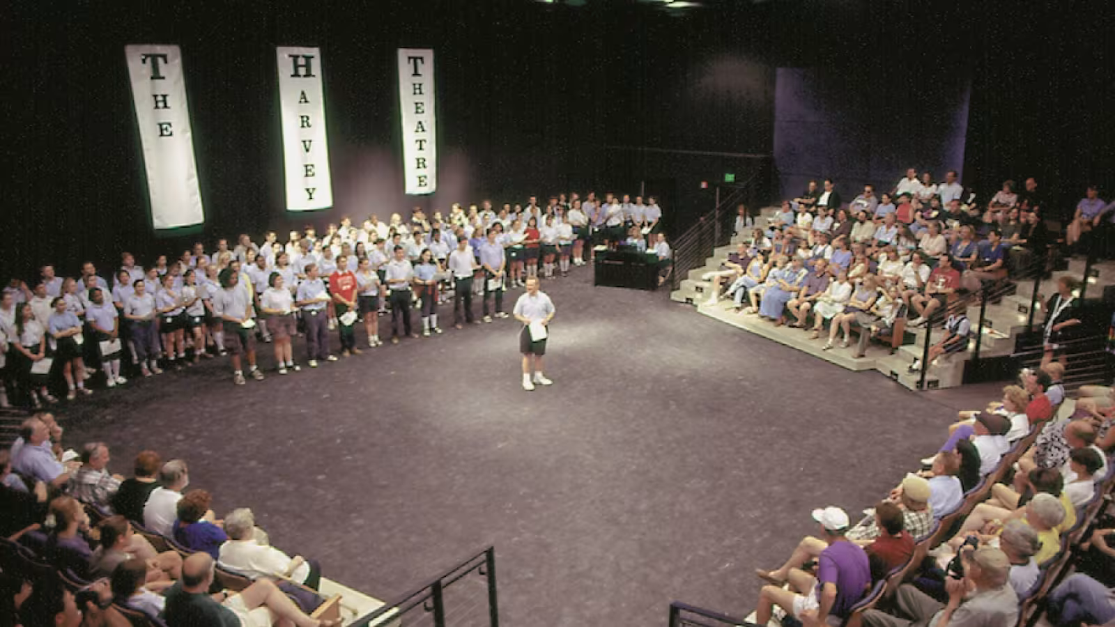 A theater with "THE HARVEY THEATRE" banners, audience on tiered seating, and people standing on the left.