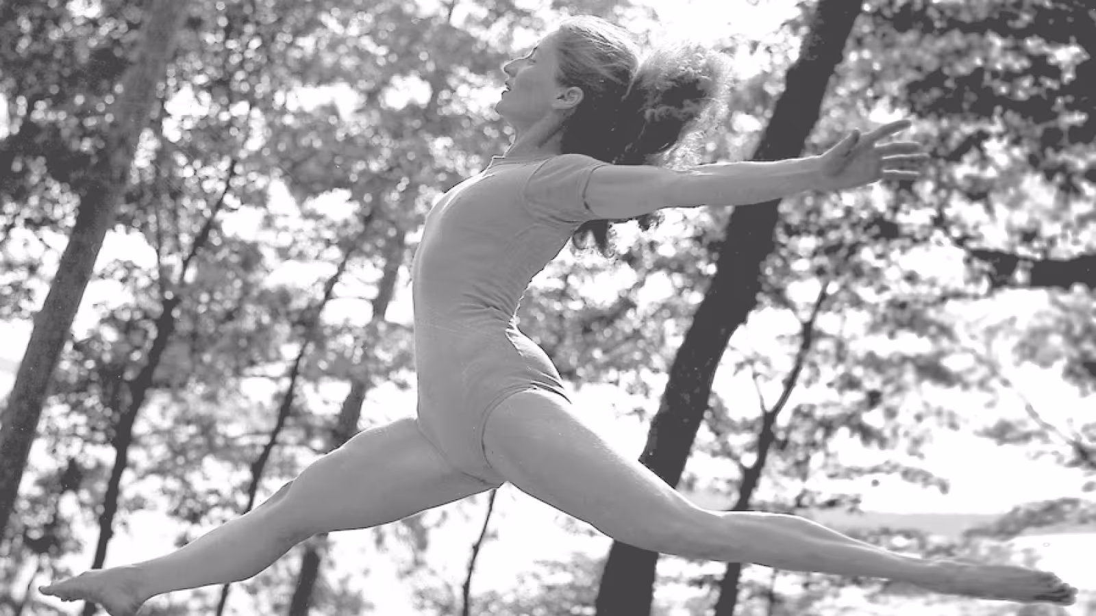 A dancer in mid-leap performing a grand jeté, positioned horizontally against a backdrop of blurred trees.