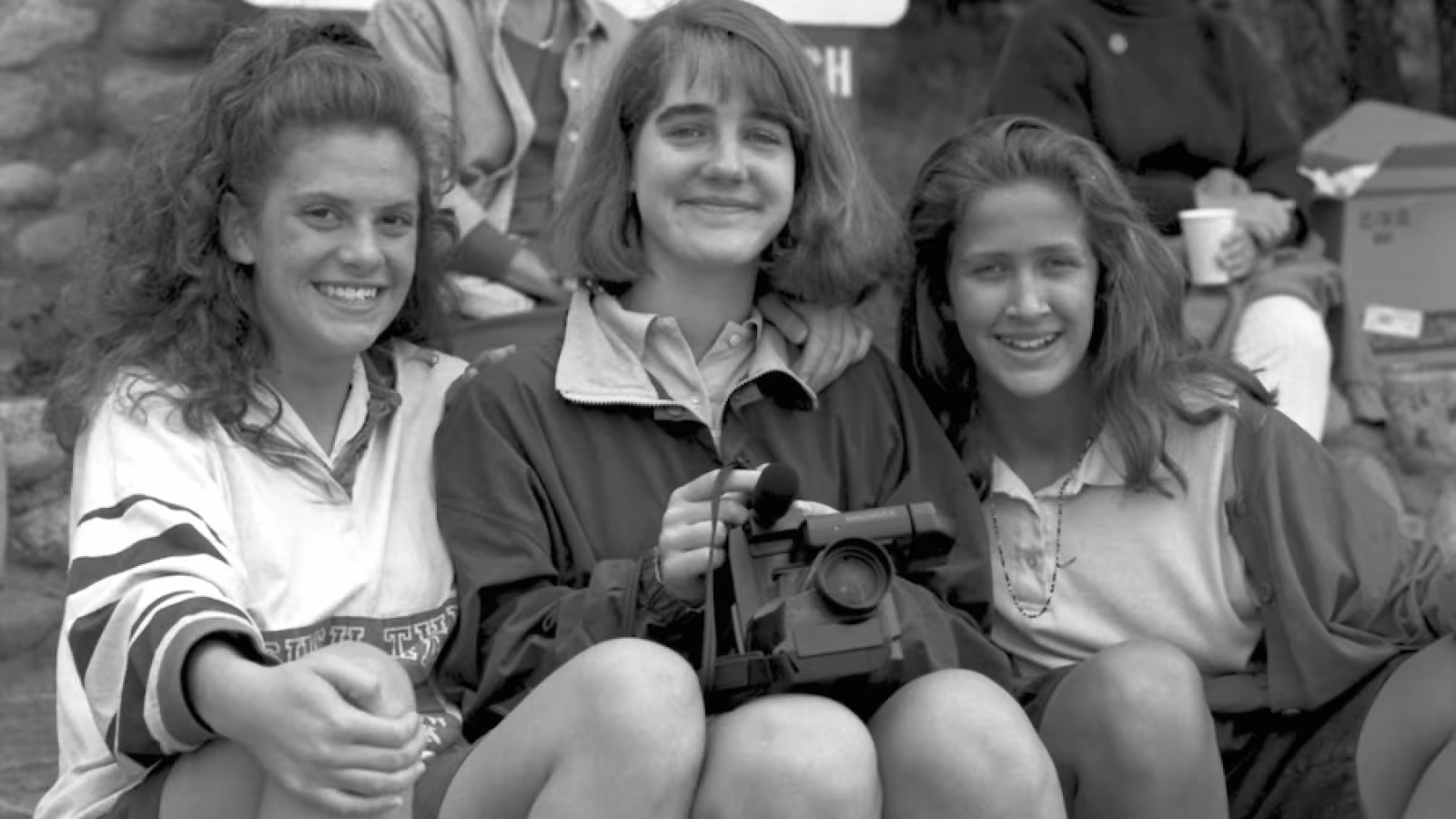 Three young women sitting and smiling with one holding a video camera. 