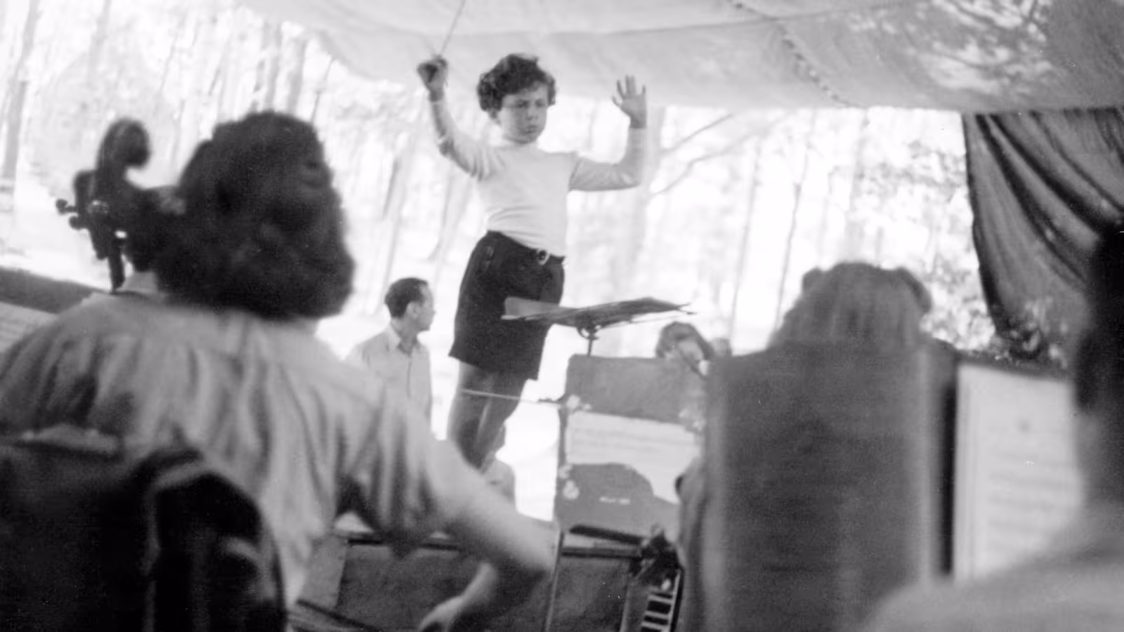 Black and white photo of a child conducting an orchestra outdoors under a canopy.