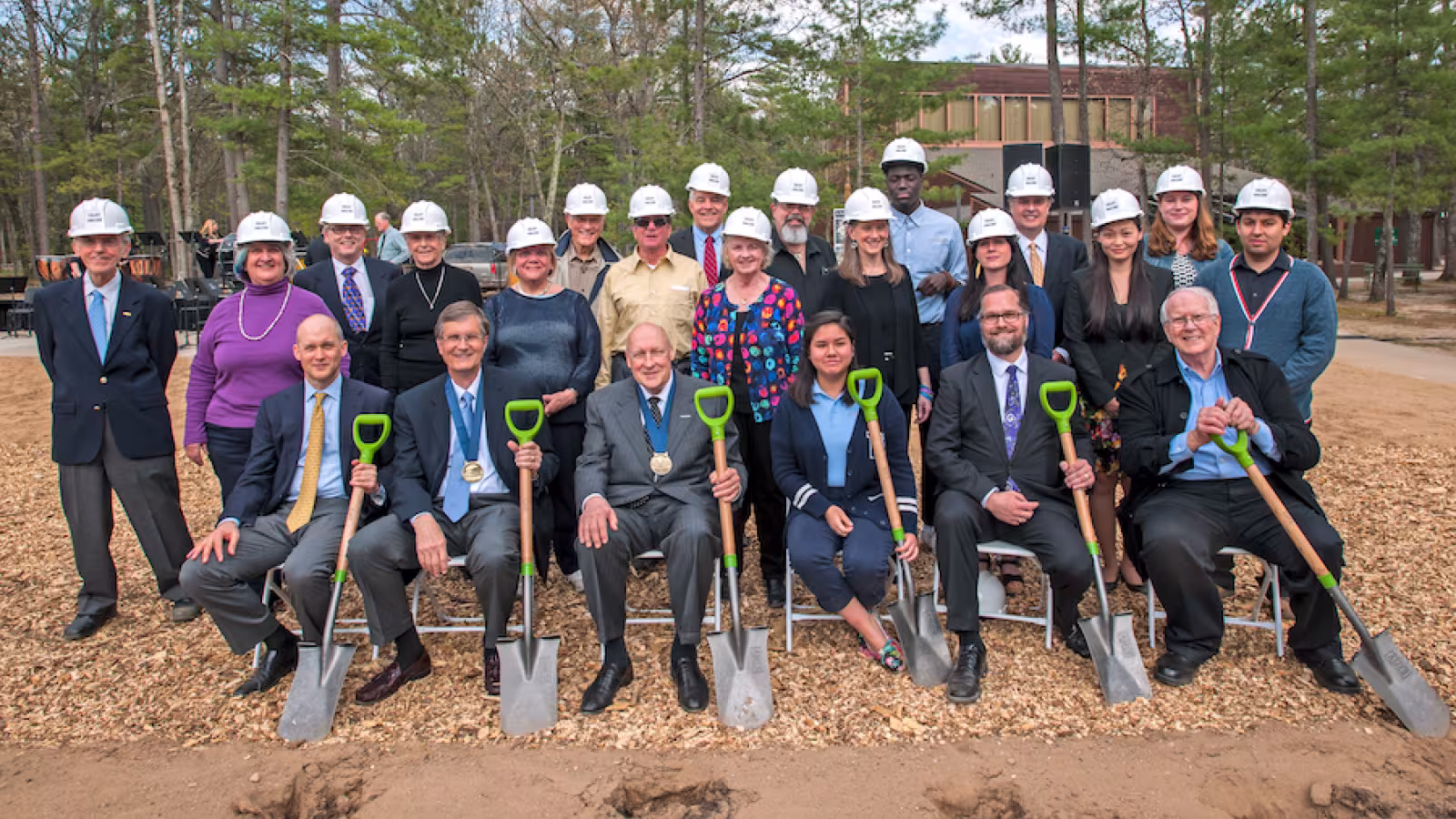 Group photo at a groundbreaking ceremony with people in formal attire and construction helmets, holding shovels.