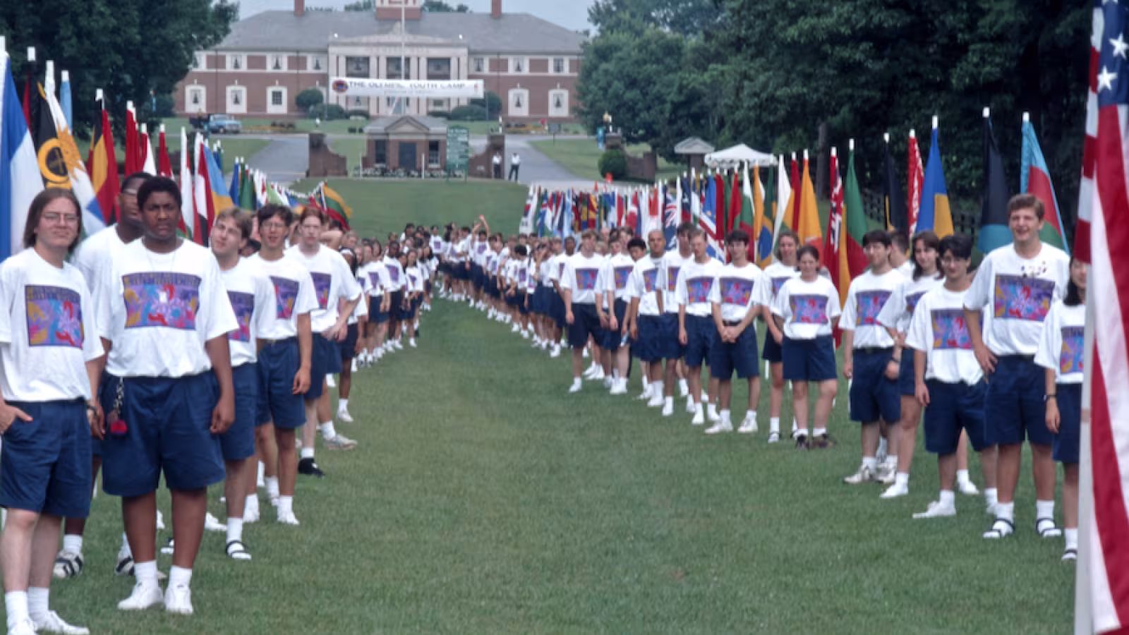 A large group of young people stands in two lines on a lawn, surrounded by international flags. 
