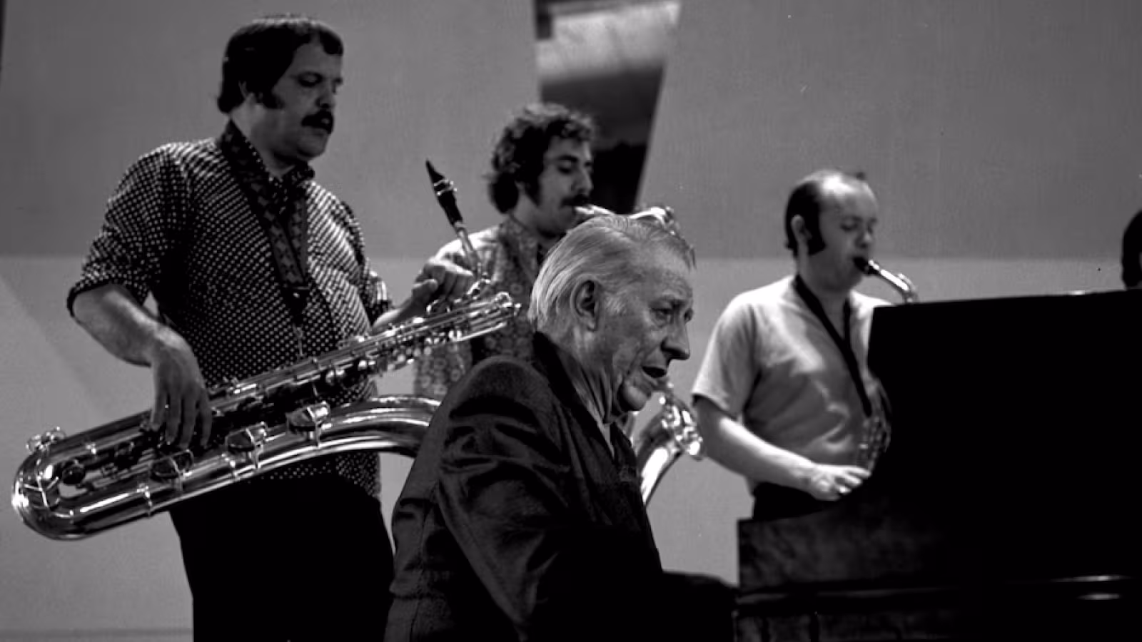 Black and white photo of Stan Kenton at the piano, backed by three musicians playing saxophones. 