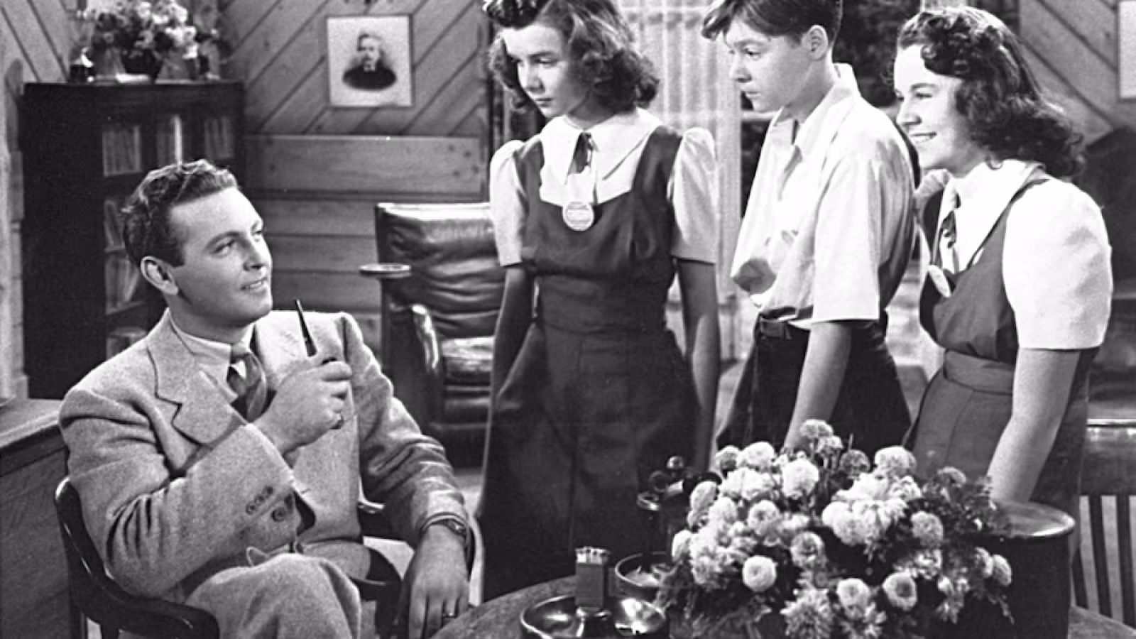 A man in a suit with a pipe talks to three young individuals in uniforms inside a room with wood-paneled walls and a table with flowers.