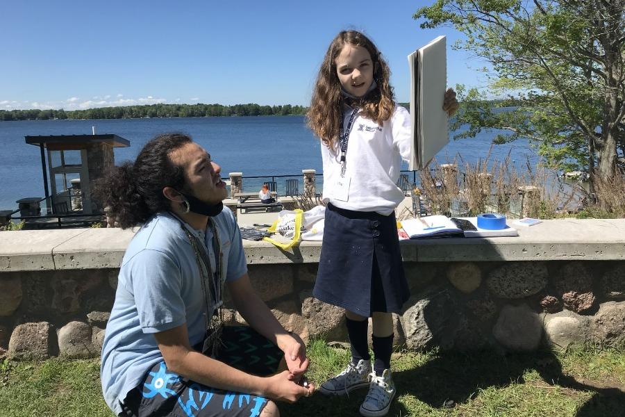 A junior Visual Arts Exploration student discusses her work with instructor and multidisciplinary artist Jamie John (IAA 15-19) on the shores of Green Lake.