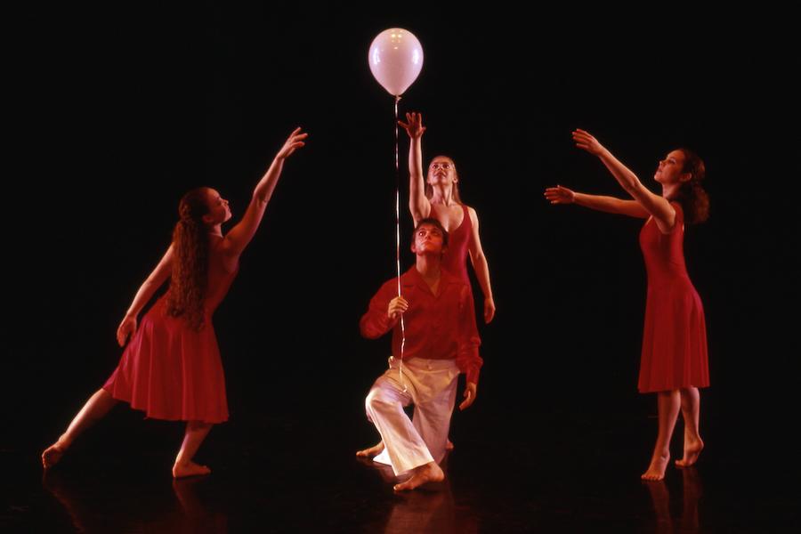 Three dancers in red stretch toward a white balloon, held aloft by a fourth dancer kneeling in the group's center. 