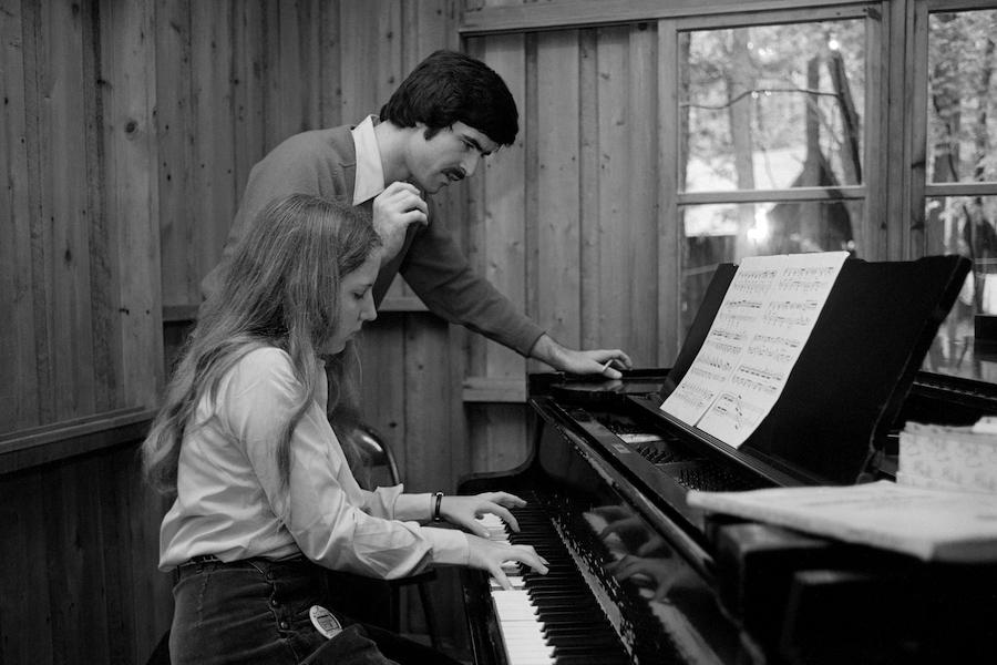 A male in his late 20s with dark hair and a mustache stands next to a female student playing piano.