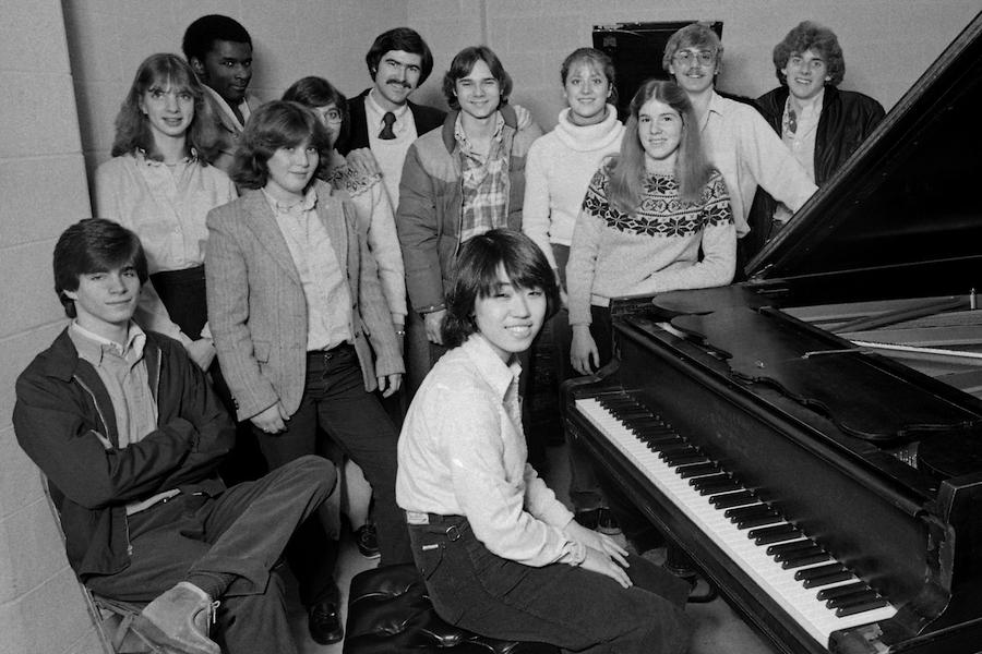 A dark-haired in his 30s poses next to a piano with 11 high school students smiling at the camera. male with a m ustacheMichael Coonrod with students from his 1981-82 Interlochen Arts Academy piano studio.