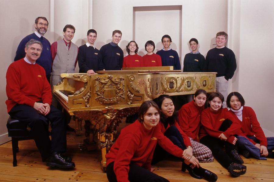 A group of piano students and teachers n blue and red sweater gather around a highly ornate and guided piano. 