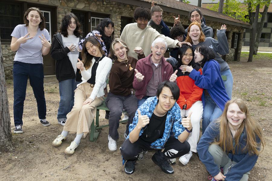 A grey-haired male in his 70s is surrounded by 14 high school students making silly faces and gestures. 