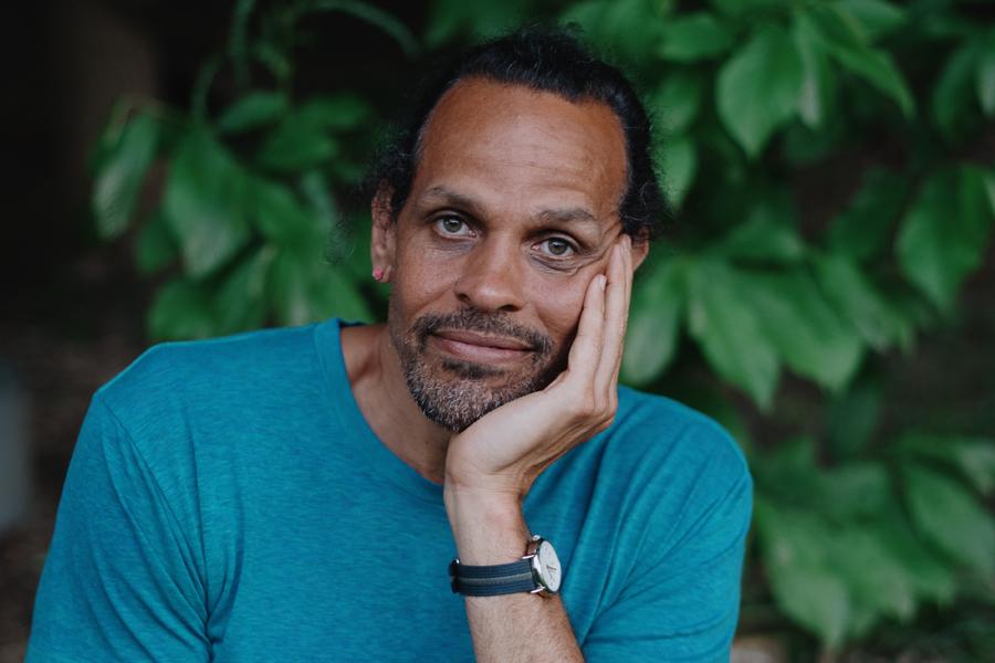 A casual portrait of Ross Gay with his hand holding his head. He wears a teal t shirt and is standing in front of a leafy bush.