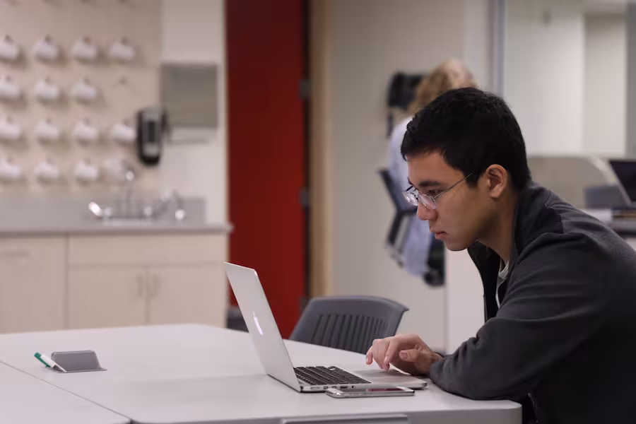 A male student with glasses, wearing a gray shirt, sits at a table and looks at his laptop screen. 
