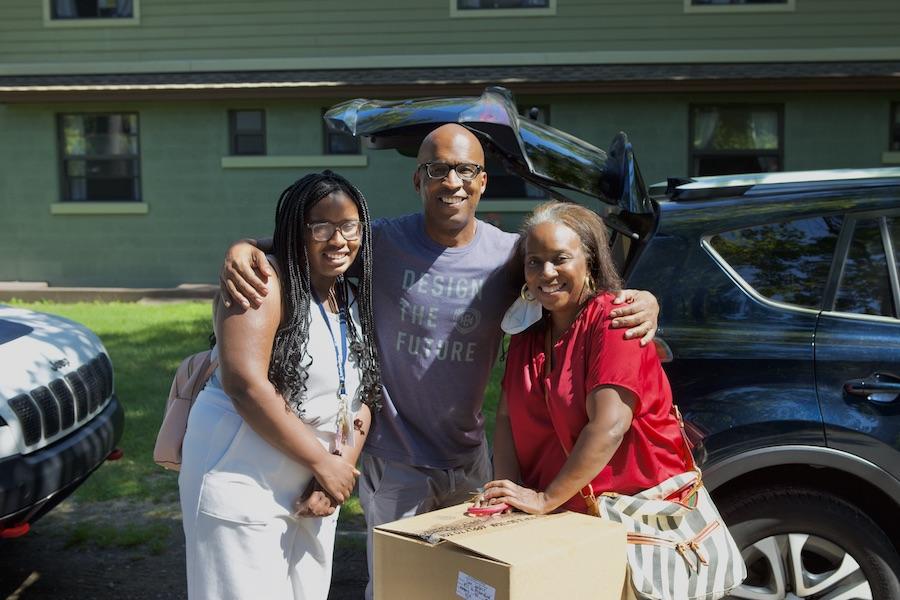 A dark-skinned female student poses in front of a car with her parents. They are in the middle of unloading cardboard boxes for move-in day.