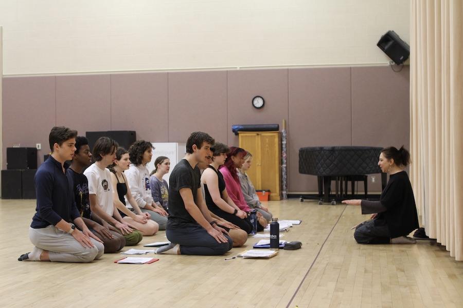 About twelve students kneel in two rows on a wooden floor, facing their teacher.