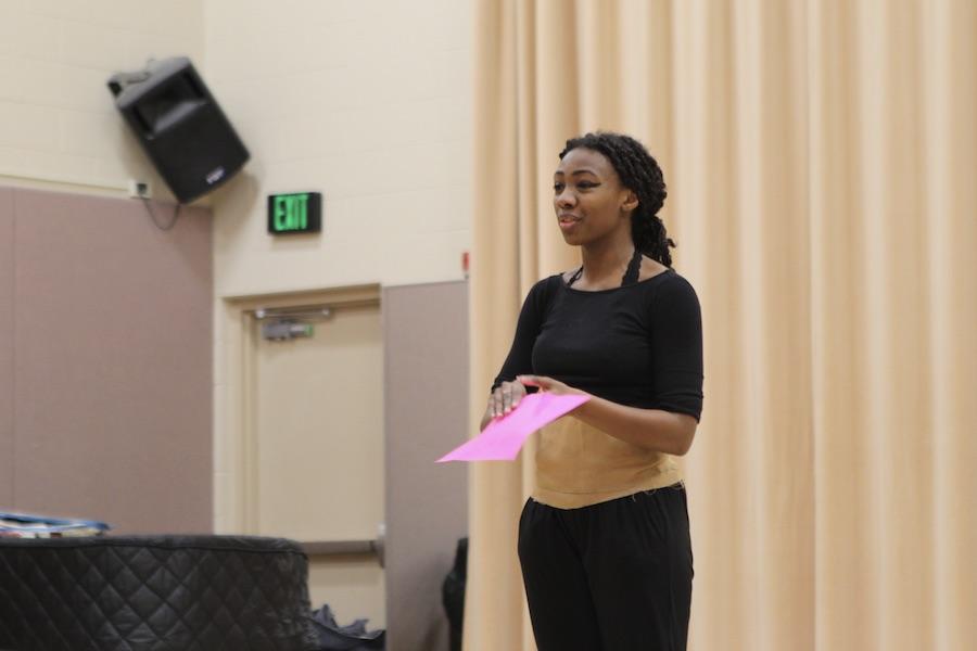 A dark-skinned student holds a piece of pink paper and speaks to an audience behind the camera.