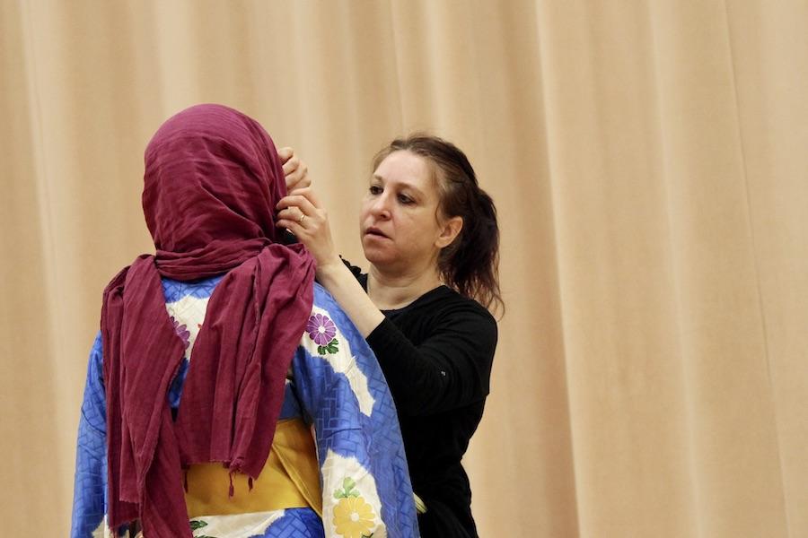 A woman wearing black adjusts the magenta scarf of a student, dressed in a blue kimono, who stands with her back to the viewer.