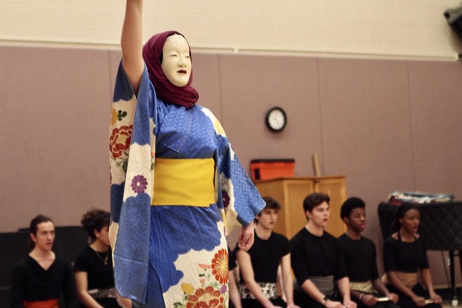 A student wearing a white Noh mask, magenta headscarf, and blue kimono raises a fan in front of a row of students who are dressed in black, kneeling on the floor.