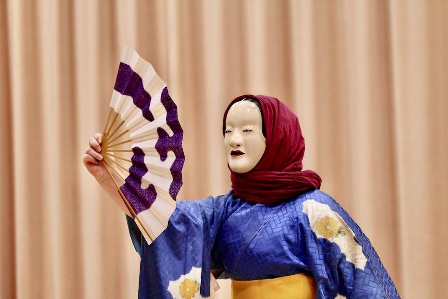 A student wearing a white Noh mask, magenta headscarf, and blue kimono raises a white fan with purple blotches.