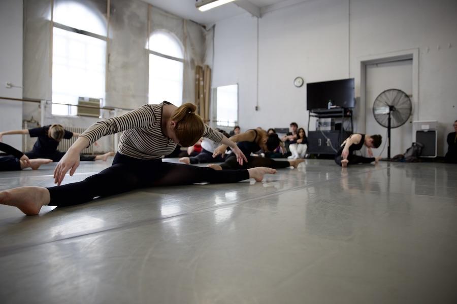 Students practice stretching in a dance studio with natural light.