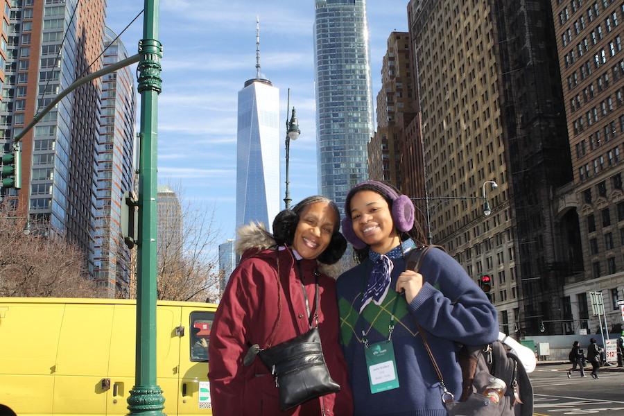 A student wearing a blue coat poses with her mother, who wears a red coat, in front of skyscrapers.