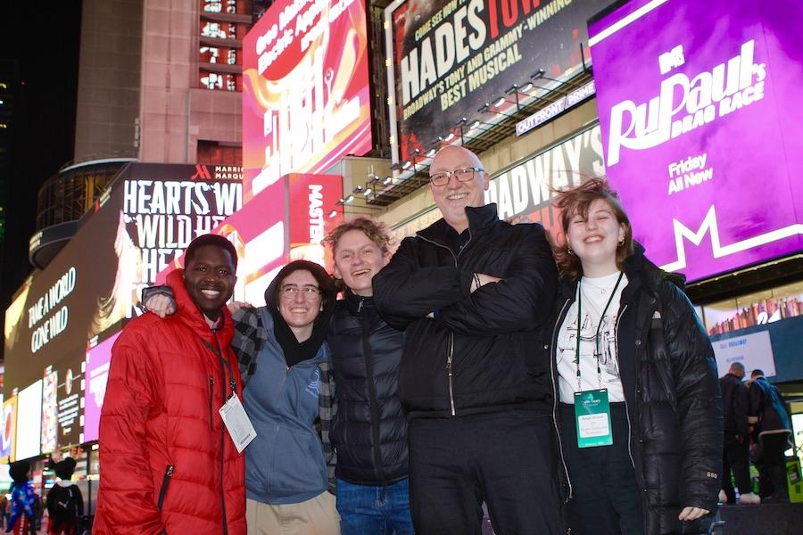 Four students and an instructor pose in front of neon signs at night in Times Square, New York.