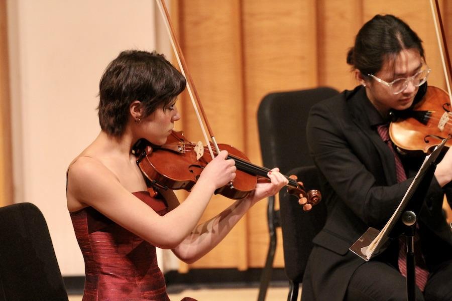 A student in a red dress plays violin in a warm wood concert hall.