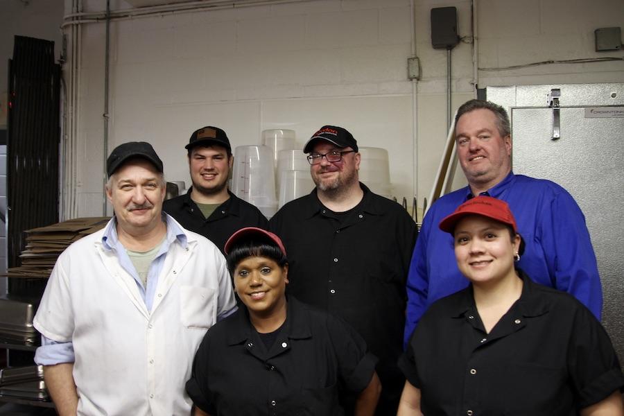A group of people dressed in food service uniforms pose together in front of an industrial fridge.