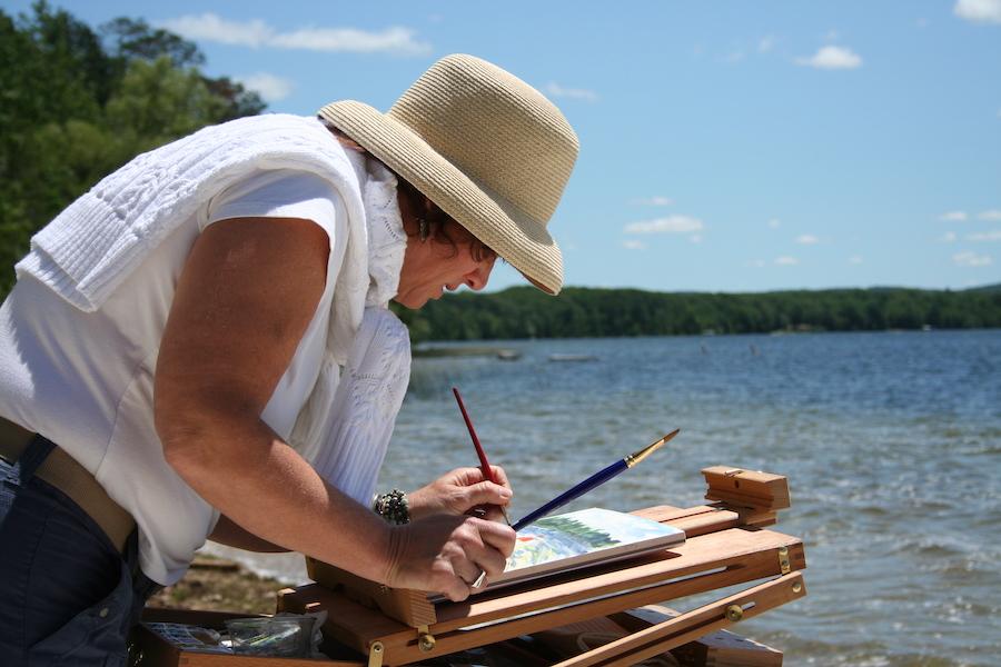 A woman paints on a canvas outside along the water. 