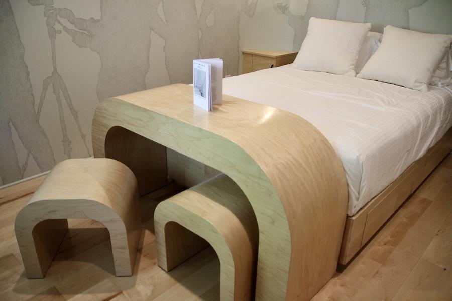 The image shows a bed with a curved desk and matching chairs made out of light-colored wood at the foot.