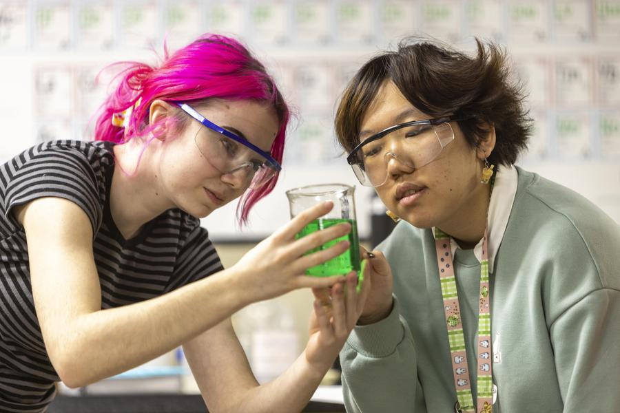 Two students look at a glass beaker filled with green fluid.