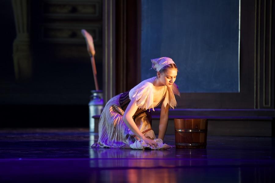 A dancer dressed in a ragged dress uses a cloth to scrub the floor.