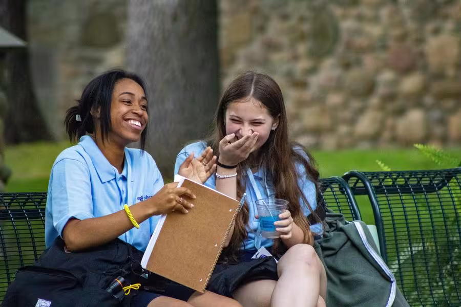 Two students in uniform smile while sitting on benches outdoors.