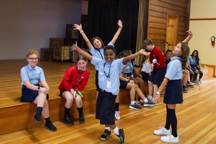 A group of students smile and wave their arms while sitting on or standing near a wooden stage.