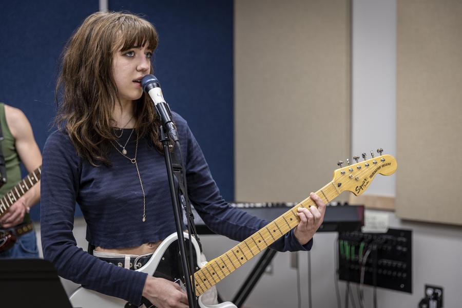 A dark-haired student sings into a microphone and strums an electric guitar.