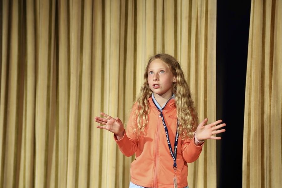 A student with long blonde hair wearing an orange shirt stands in front of a curtain to deliver a monologue.