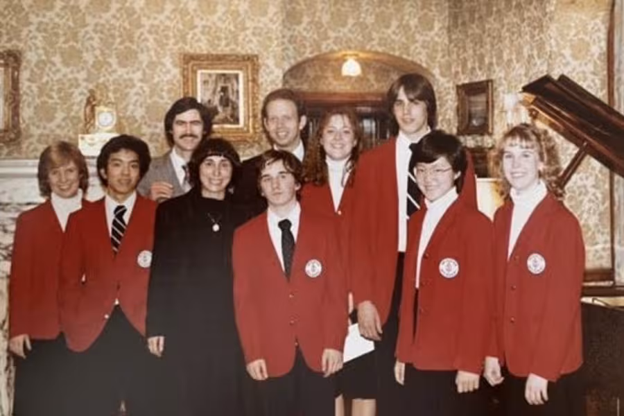 A group of students and teachers stand and smile in a room with gold wallpaper.