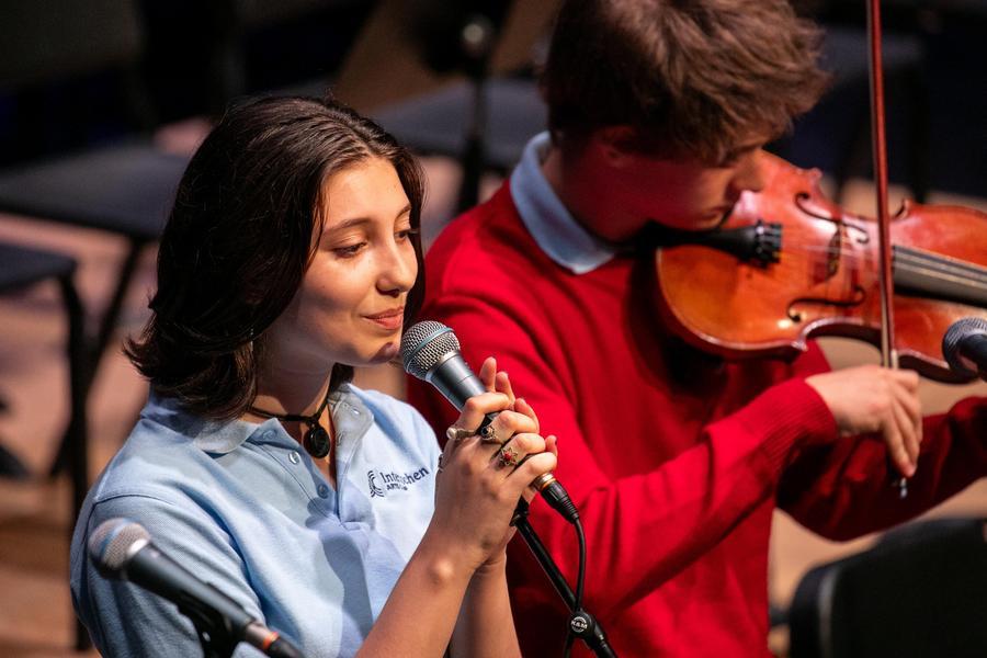 Two students in Interlochen attire—one with a microphone, and the other with a violin—perform on stage.