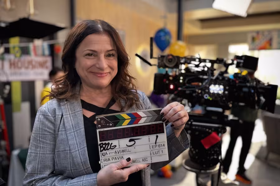 A smiling director poses with a clapperboard in front of camera equipment.