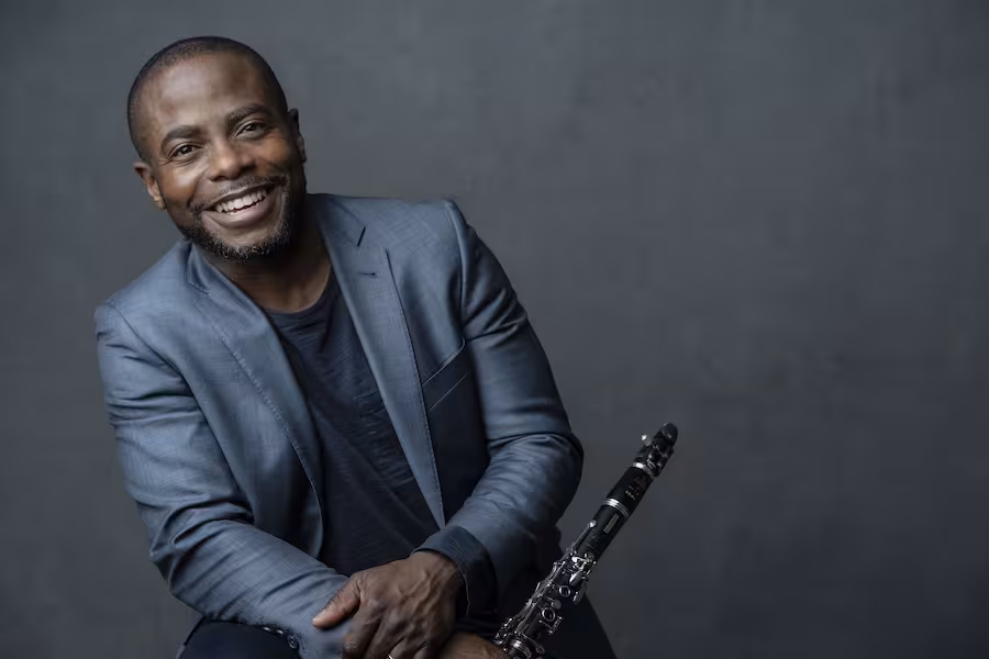 Anthony McGill stands in front of a grey background and smiles while holding his clarinet. 